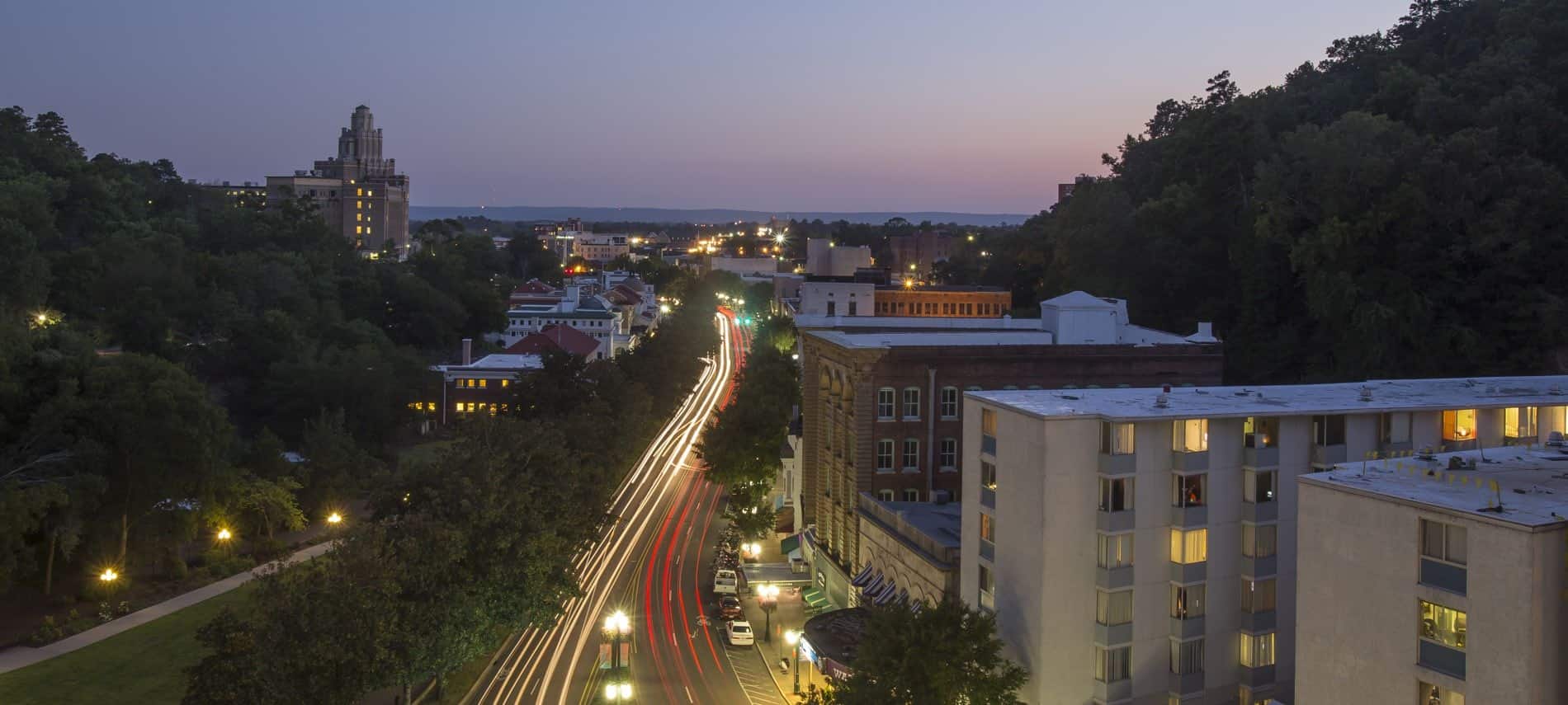 Overview of a city at dusk with a slightly winding road, buildings with lights, and many green trees