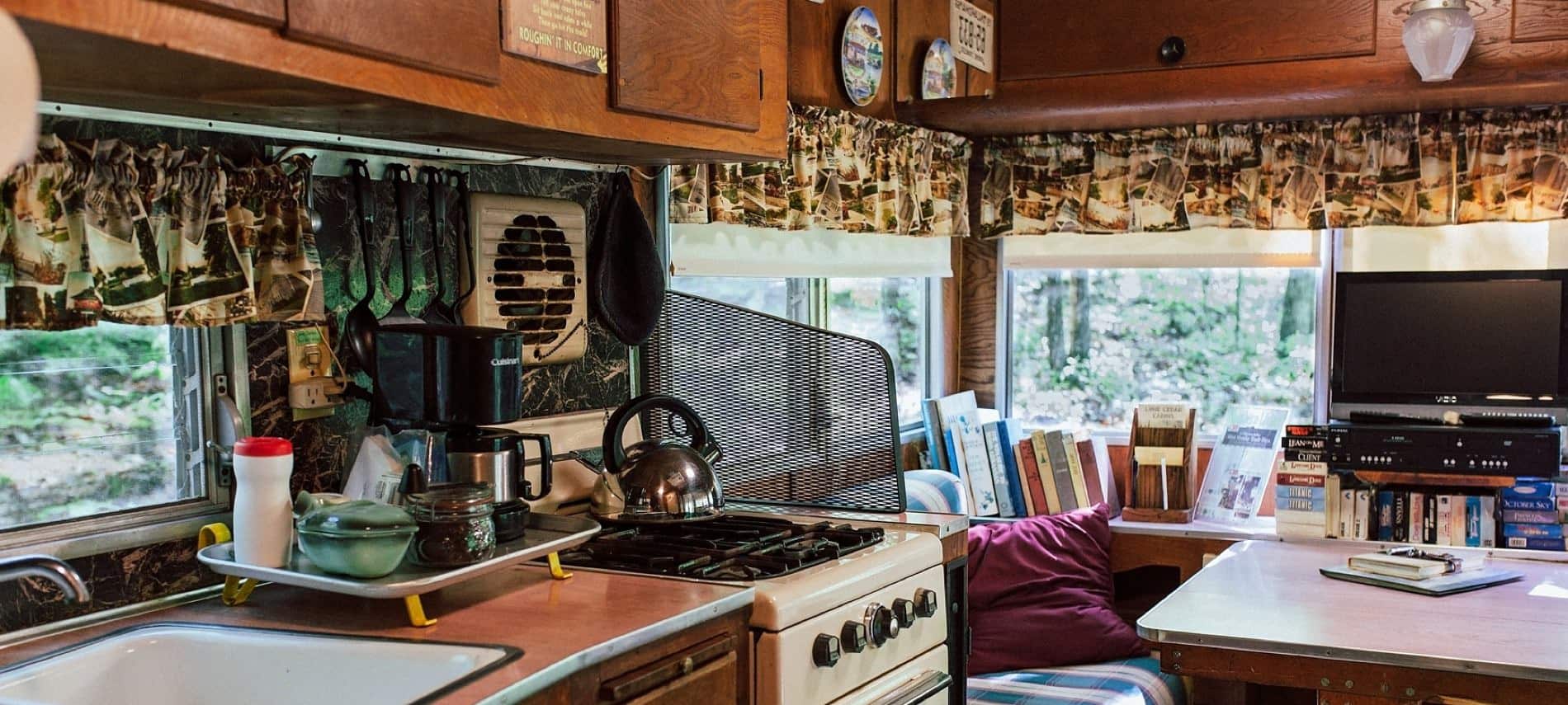 Inside of cabin with sink, range, and table with built-in seats, shelf of books and small flat screen TV