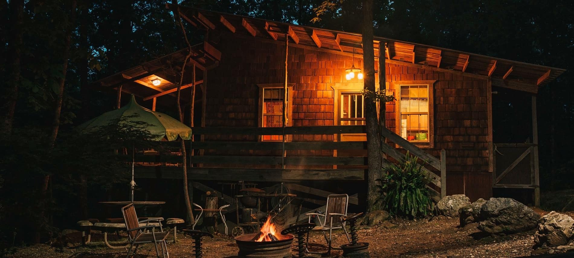 Cabin exterior at night with round patio umbrella table and chairs around a warm glowing fire pit