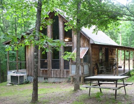 Exterior view of wooden cabin with gable roof, two side covered porches, a picnic table, surrounded by green leafy trees