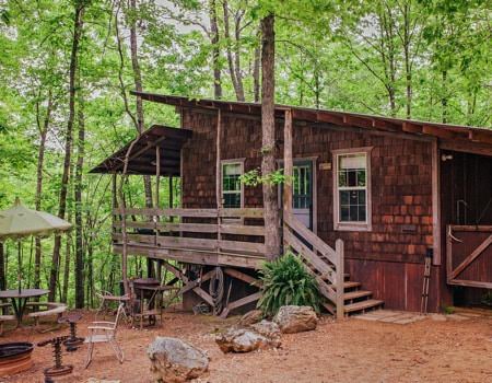 Exterior of reddish brown cabin with side covered porches, round picnic table with umbrella and fire pit surrounded by trees