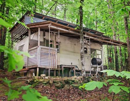 Exterior of cabin and charcoal grill in the woods surrounded by trees with leafy green leaves