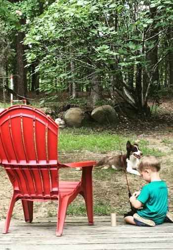 Little boy with fishing rod sitting on a wooden deck by a red chair and a brown and white dog